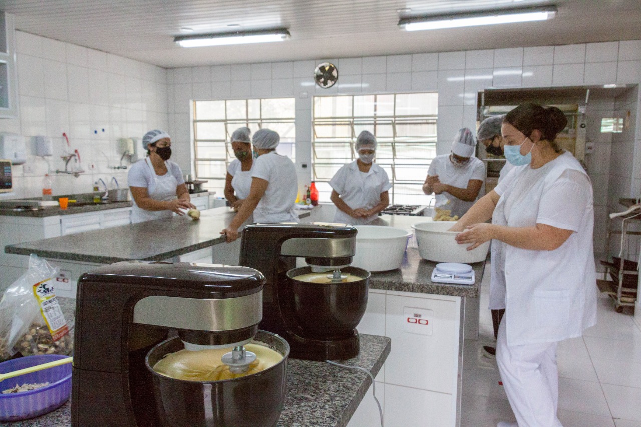 Cozinha com sete mulheres usando equipamentos de proteção individuais preparam refeições em bancadas, com auxílio de batedeiras. 