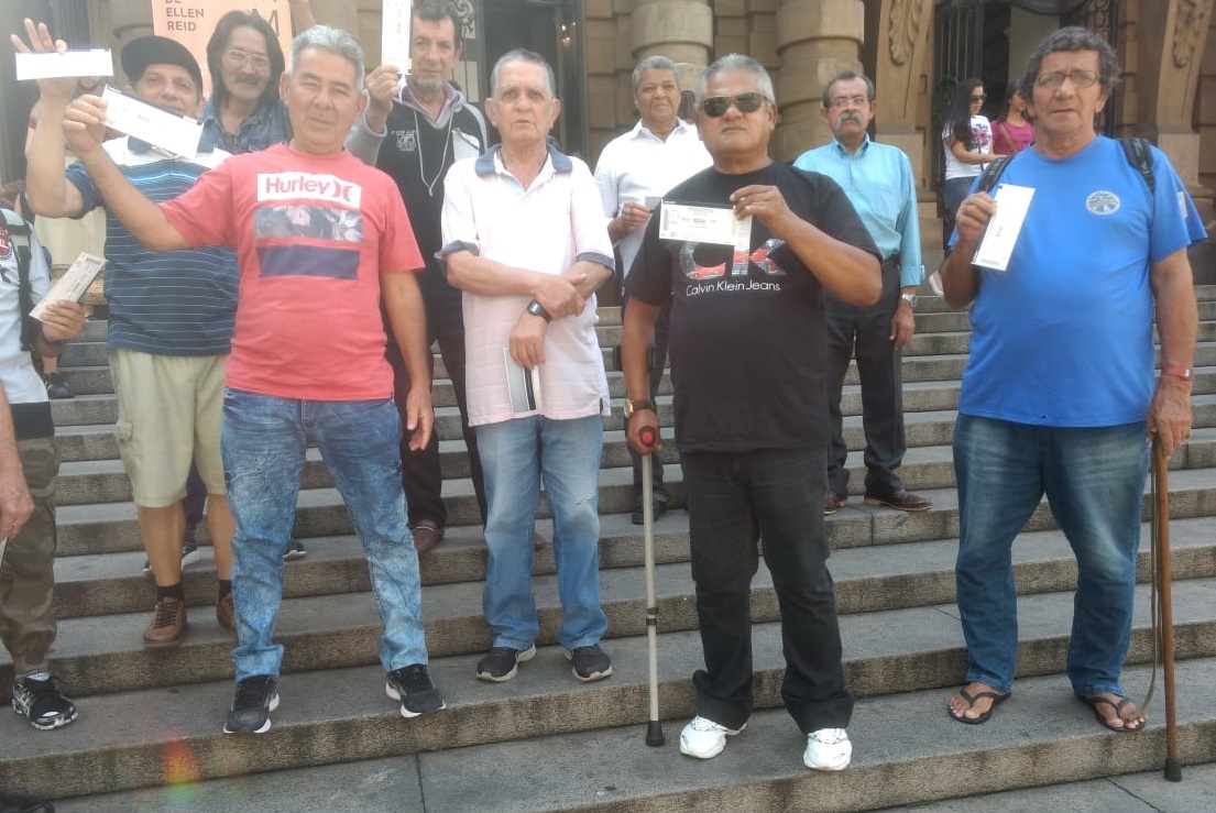 #PraCegoVer:Foto 1: Nove homens em pé em frente ao Theatro Municipal segurando ingressos enquanto posam para foto.