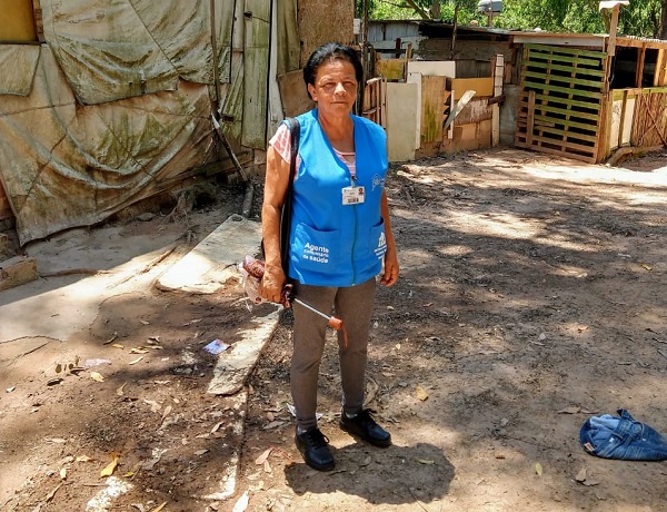 #PraCegoVer: Agente de saúde está em pé, com um guarda-chuva na mão direita e ao redor dela há casa improvisadas de madeira, com o chão de terra.