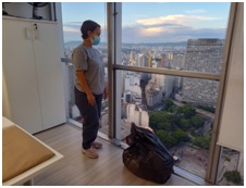Mulher de cabelos curtos, de máscara, com o saco de lixo ao seu lado, olhando a vista da cidade de São Paulo da janela do prédio. Está vestida com camiseta de uniforme de trabalho, calça jeans e tênis rosa.