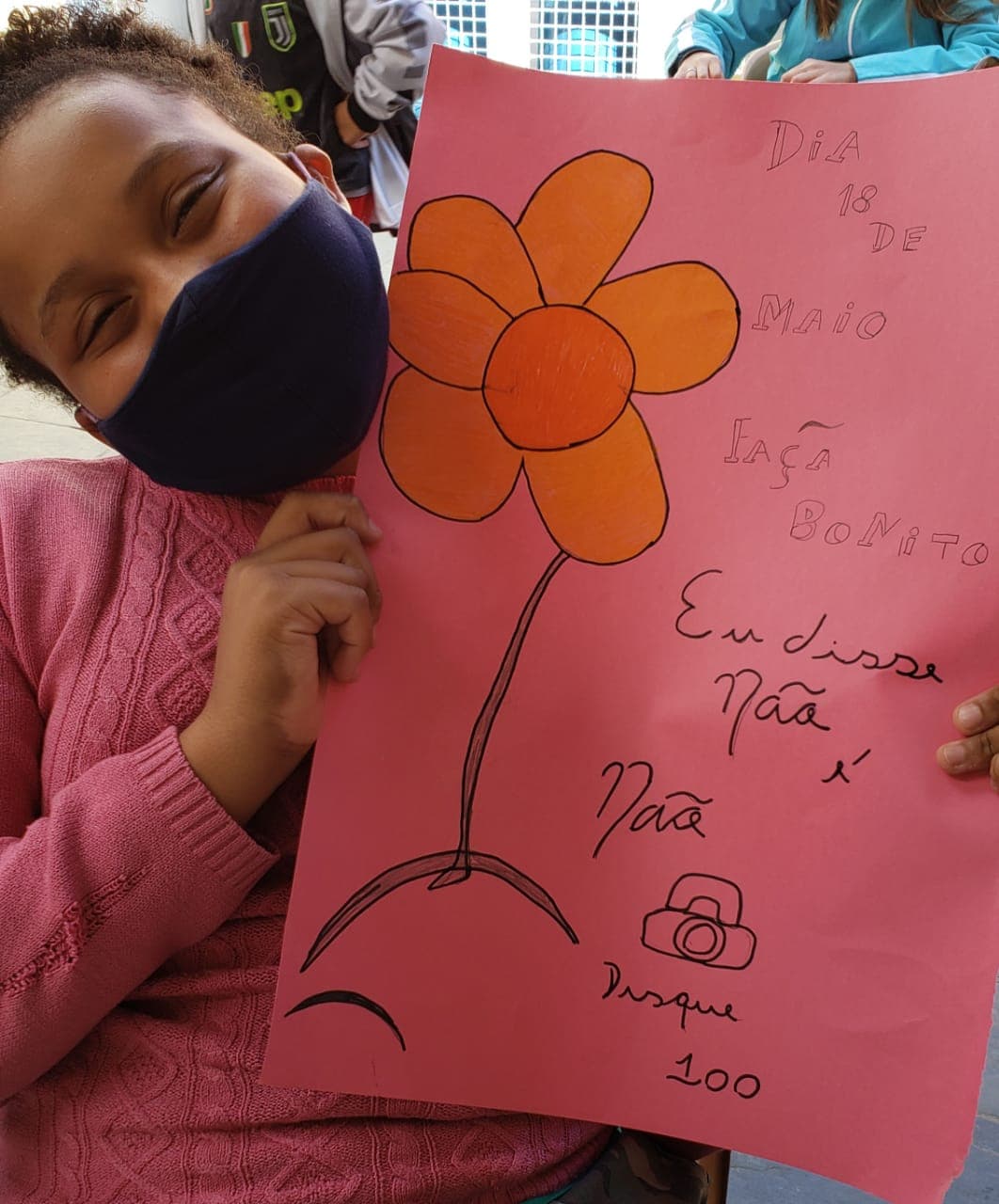 Foto com outras crianças ao fundo e na frente uma criança de mascara e blusa rosa segurando um cartaz rosa com um desenho de flor escrito de preto “Dia 18 de maio. Faça Bonito. Eu disse não é não. Disque 100”.