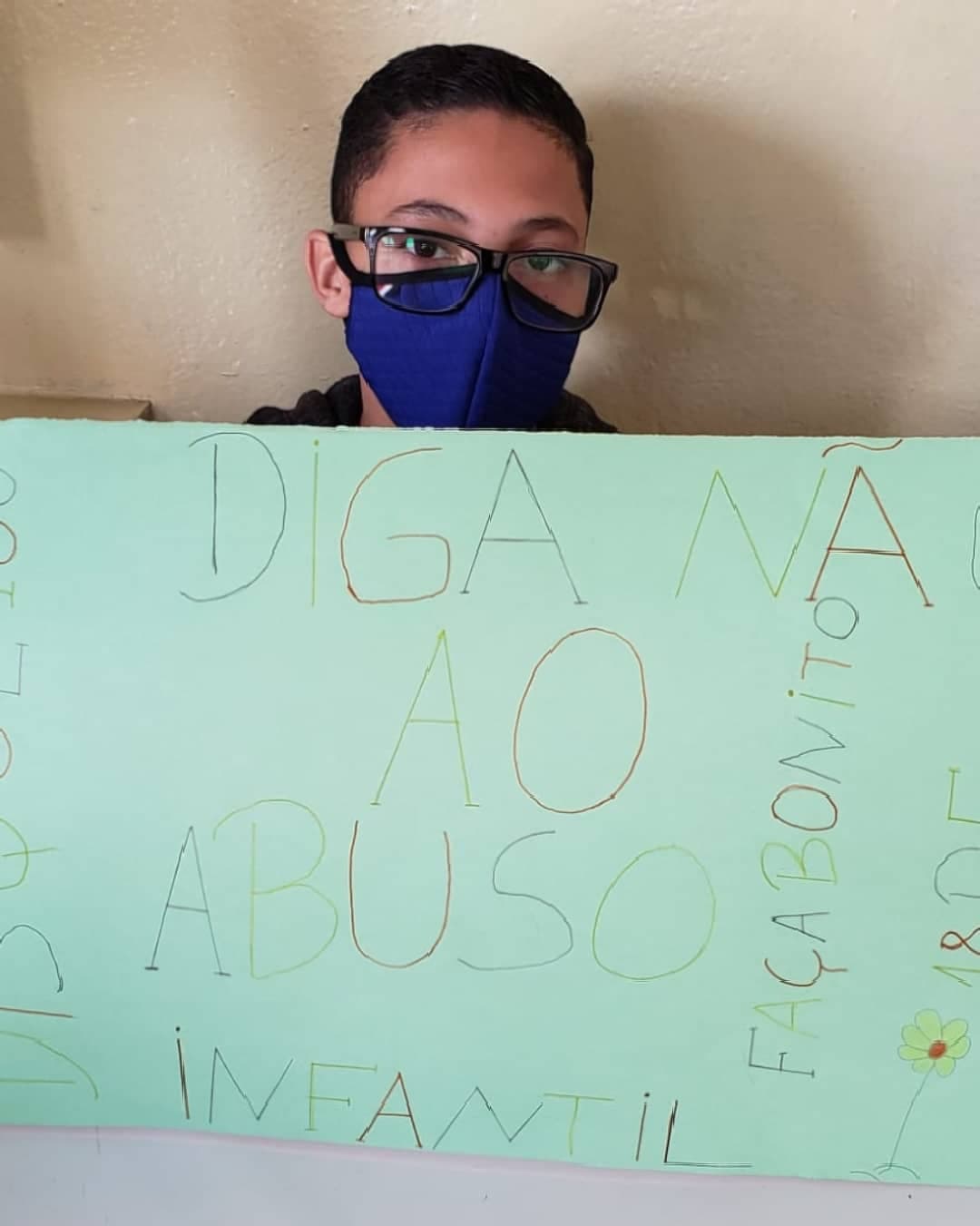 Foto com fundo creme com uma criança com mascara e óculos segurando um cartaz da cor verde escrito de varias cores “Diga não ao abuso infantil. Faça bonito. 18 de maio”.