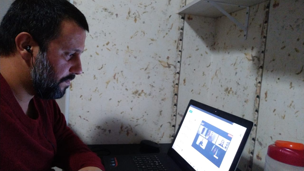 Luiz Fernando Francisquini está na lateral esquerda da imagem, de lado, vestindo uma blusa vermelha e olhando para a tela do computador. Atrás dele está uma parede de tons claros e do lado esquerdo da foto está o computador usado para a live.