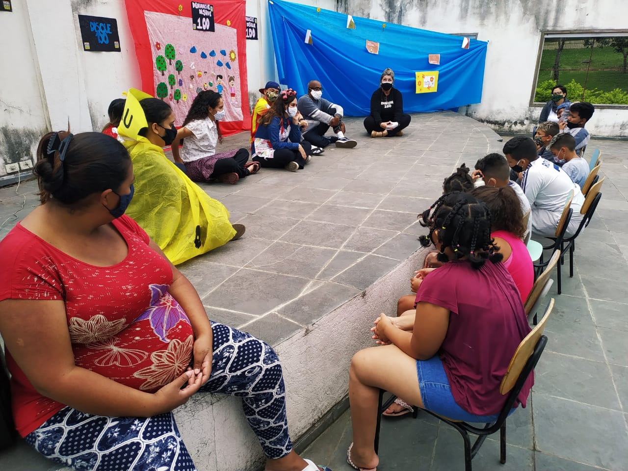 Em um espaço aberto com um palco amplo, estão outros funcionários do serviço durante a apresentação teatral cercados por crianças sentadas ao redor do palco