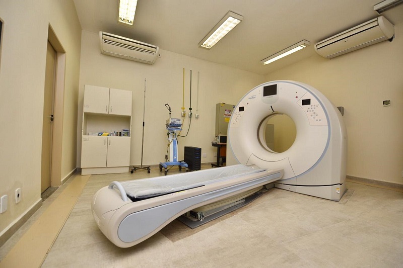#PraCegoVer: É uma sala. Ela tem tons claros, há ar condicionado, uma maca, e a máquina de tomografia. Também há um armário na cor branca.  