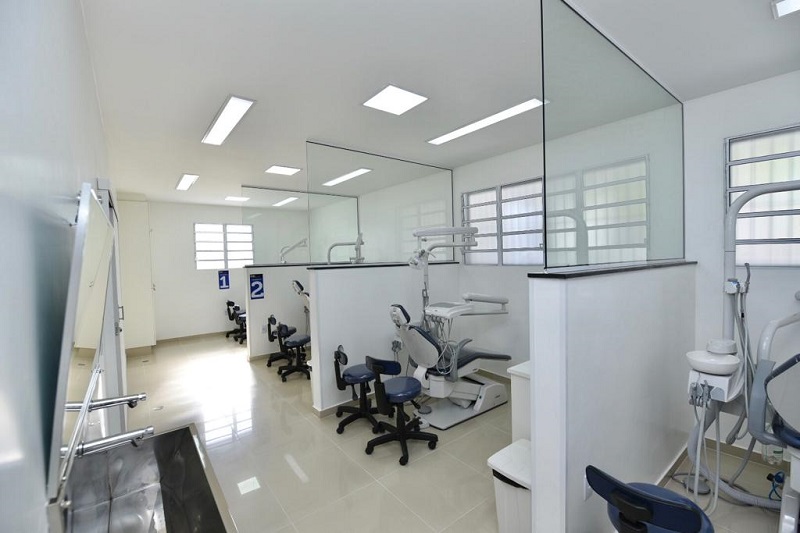 #PraCegoVer: Uma sala grande, com quatro divisórias. Em cada divisória há uma cadeira de dentista, com outros materiais odontológicos. A sala é clara, muito iluminada e com as paredes brancas.