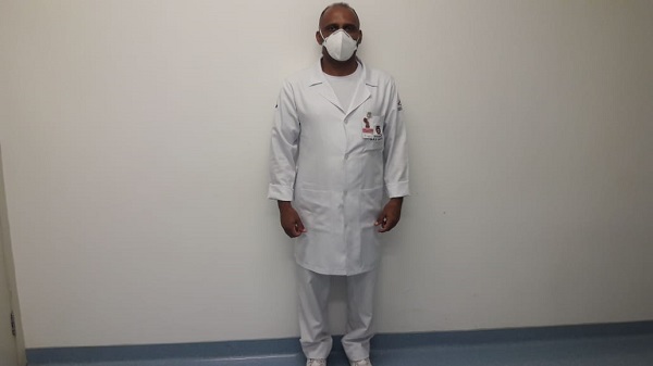 #PraCegoVer: Homem em pé, vestido todo de branco, com uniforme de enfermeiro, usando máscara. Em uma parede de fundo branco