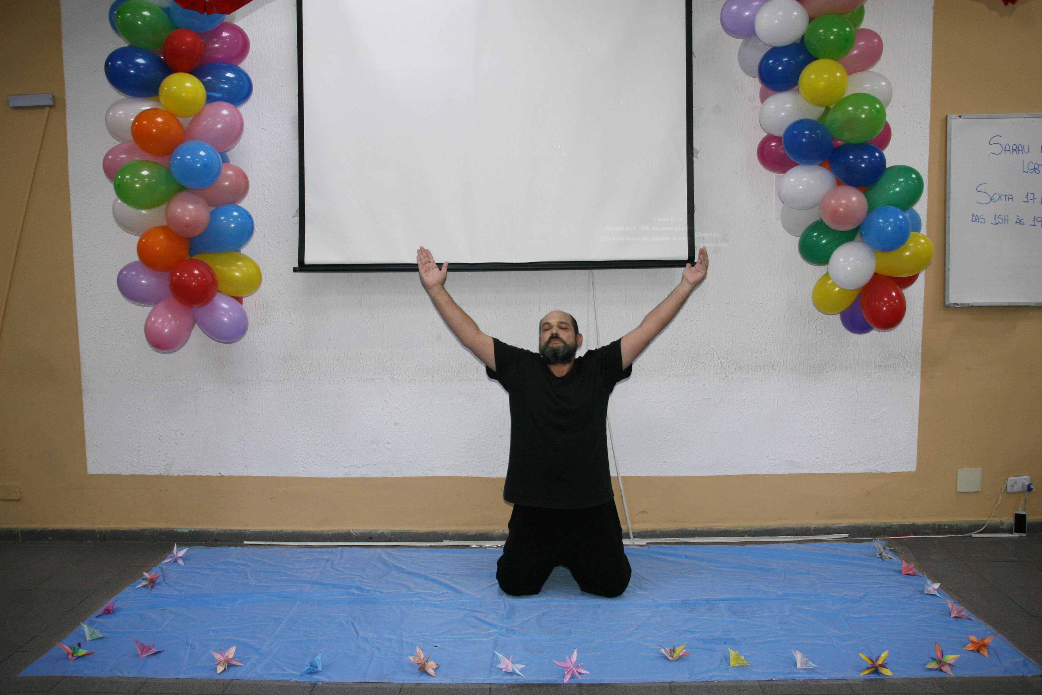 Convivente vestido de preto está com os joelhos no chão e braços para o alto em apresentação artística na sala do serviço com balões coloridos e um telão de projeção na parede ao fundo