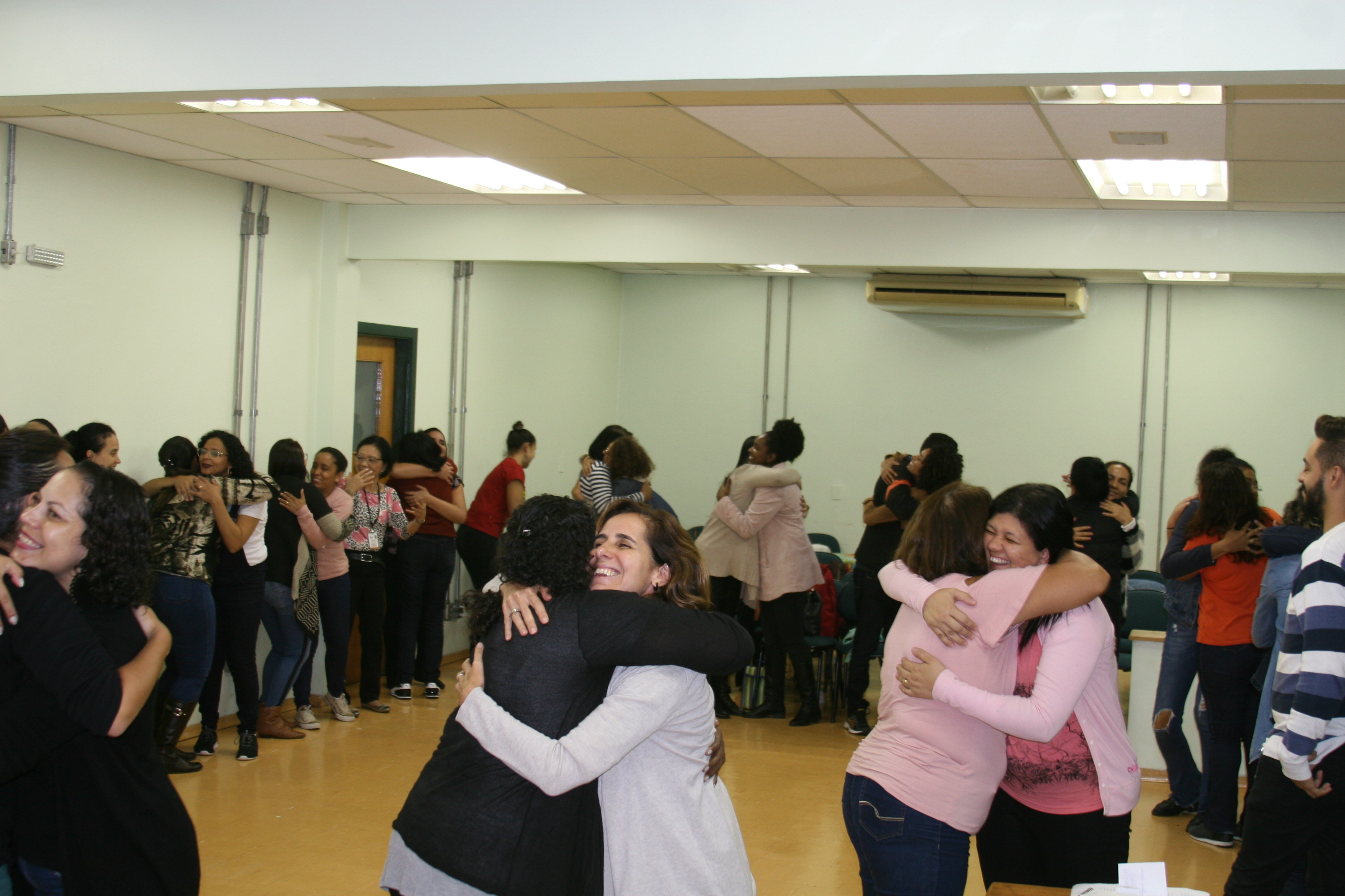 Assistentes sociais realizam dinâmica em grupo na qual se abraçam em pares formando roda enquanto sorriem