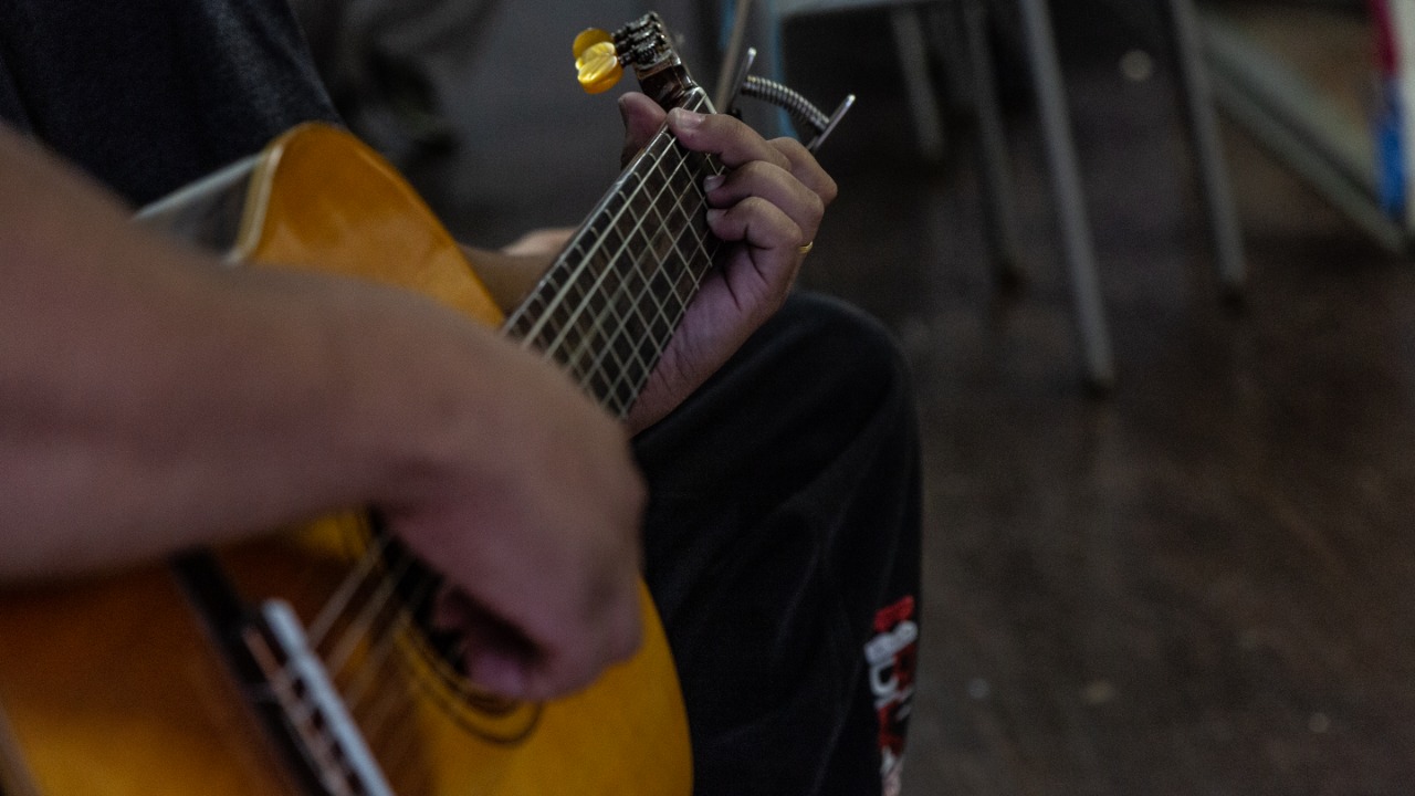 Plano detalhe de violão sendo tocado por convivente do qual se vê apenas os braços e parte da perna