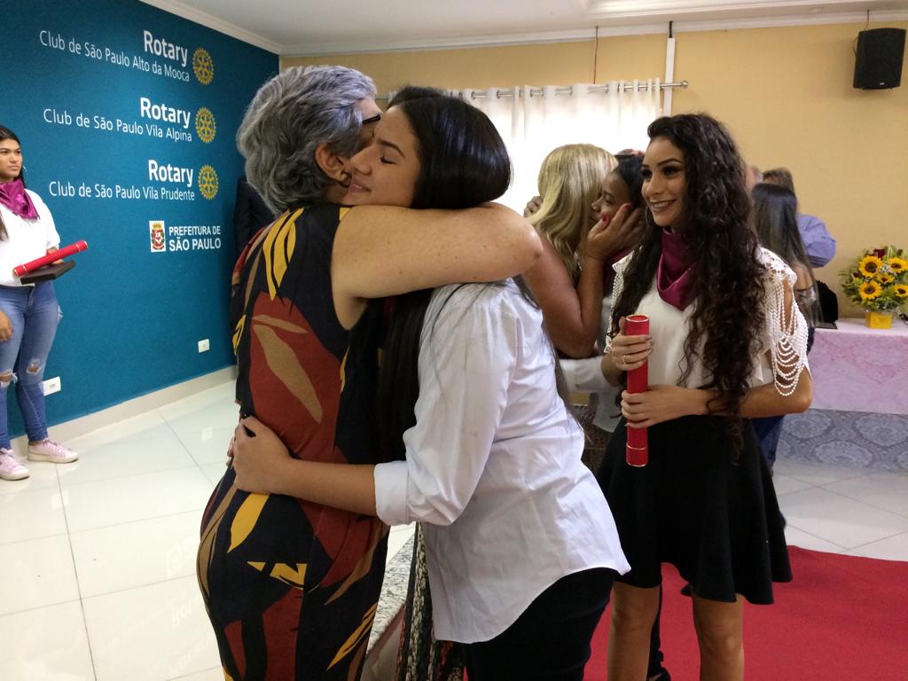 Formanda é abraçada por uma mulher enquanto em segundo plano uma outra formanda aguarda com canudo na mão e outras estão atrás em fila