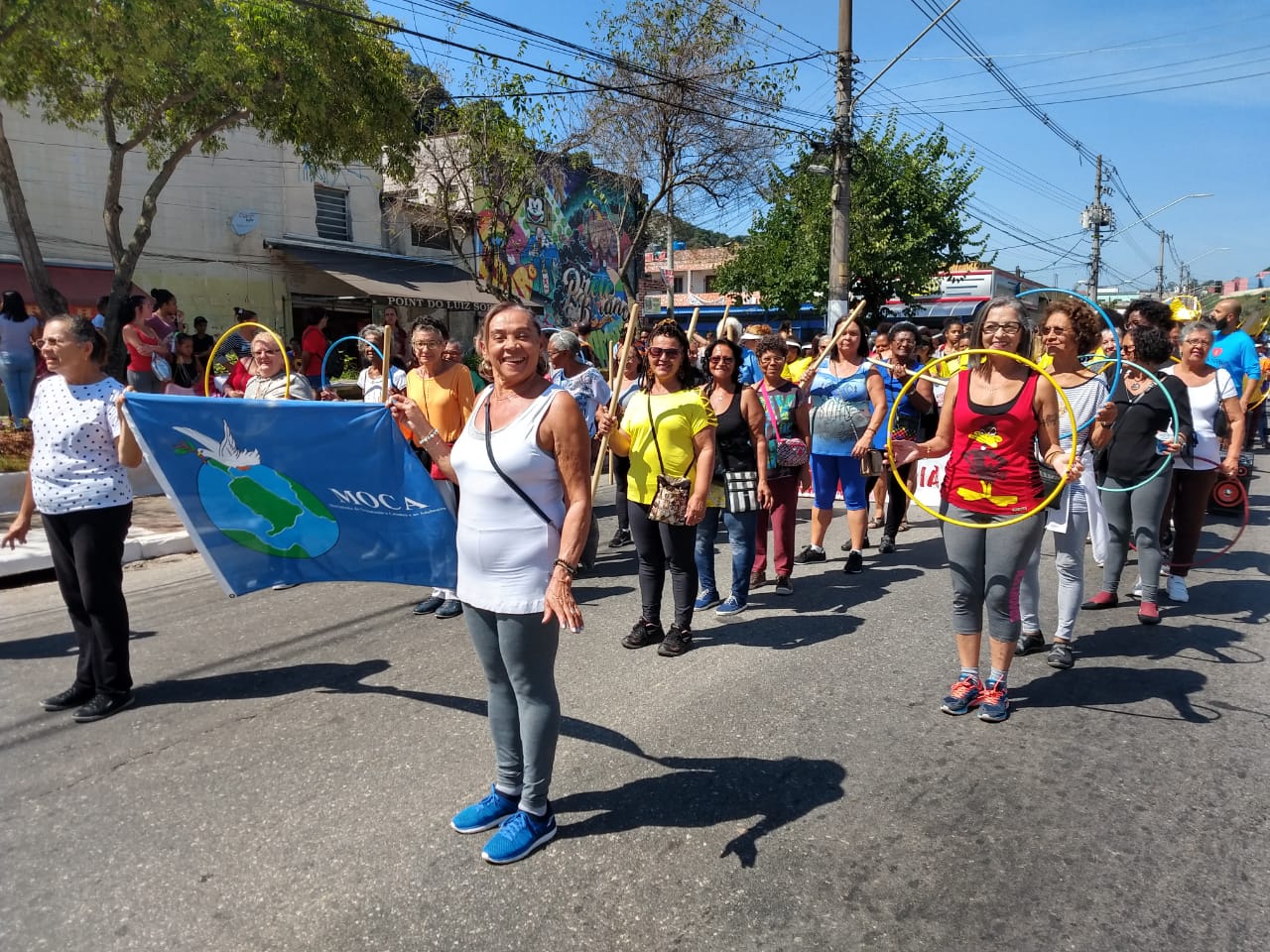 Duas senhoras conviventes do serviço segurando bandeira azul da organização gestora MOCA, enquanto outras mulheres seguem atrás no desfile com bambolês coloridos nas mãos