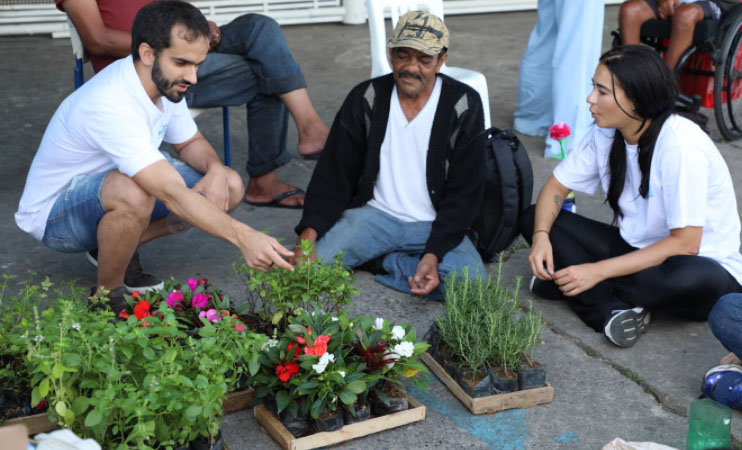 Dois voluntários e um usuário do serviço plantam mudas de plantas sentados no chão