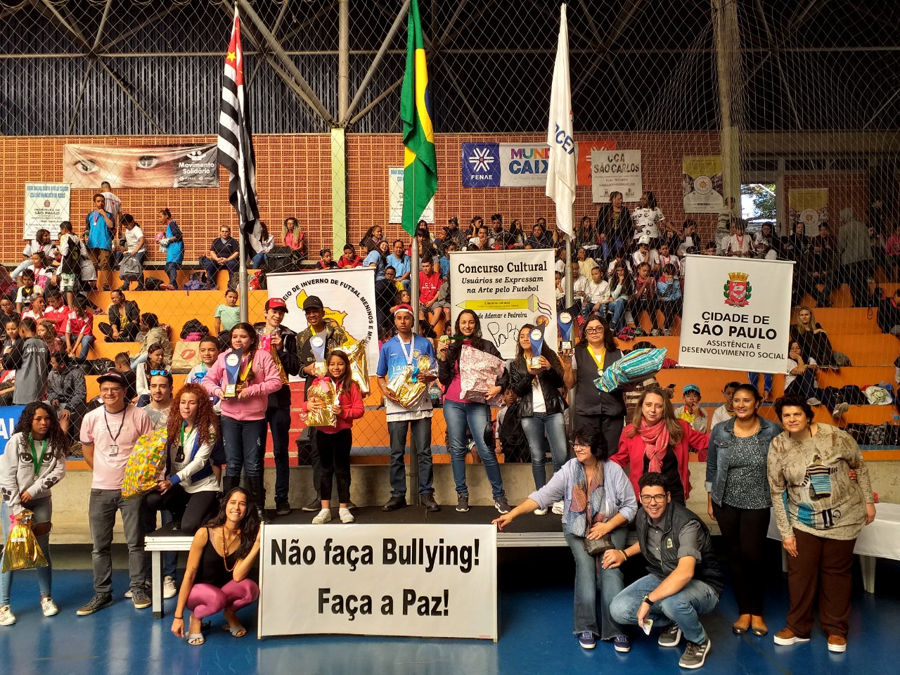 Conviventes, servidores públicos e convidados em arquibancada da arena de futsal de Cidade Ademar. Alguns sentados ao fundo e outros em pé na frente. Cartazes colados na parede de trás e um cartaz branco de letras pretas centralizado, no chão, com os dizeres não faça bullying, faça a paz
