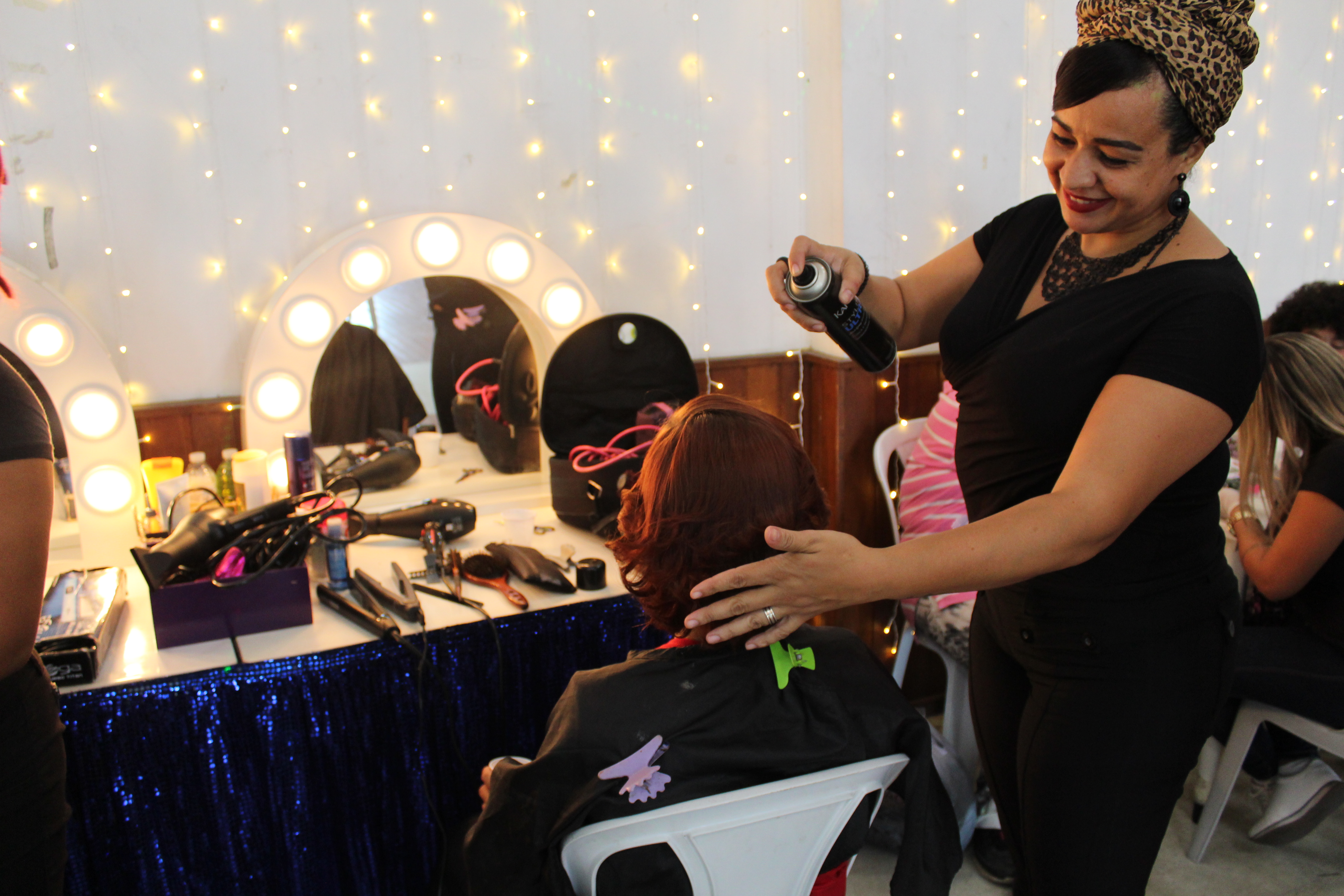 Cabeleireira com turbante de estampa de oncinha passa spray no cabelo de uma convivente sentada em frente a um espelho de salão de beleza