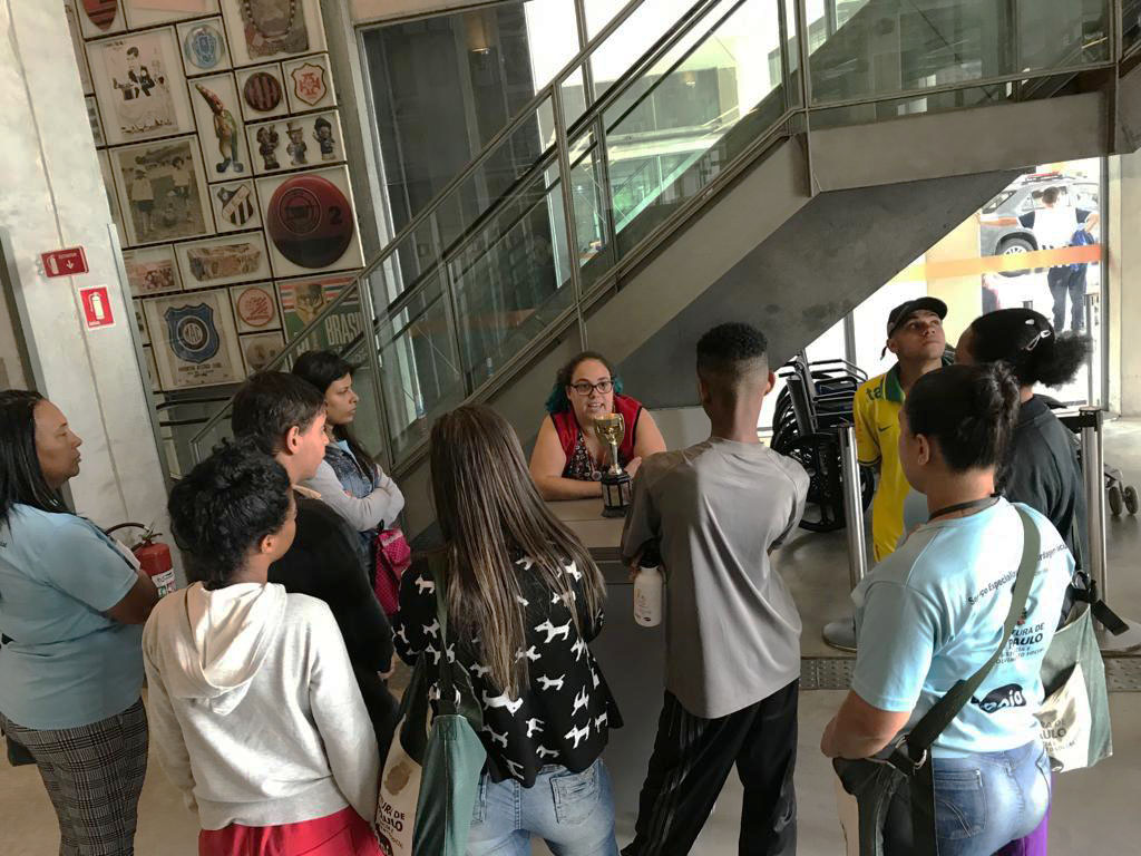 Orientadora do Museu segura taça dourada sentada atrás de uma mesa enquanto fala para plateia de jovens em pé a sua frente 