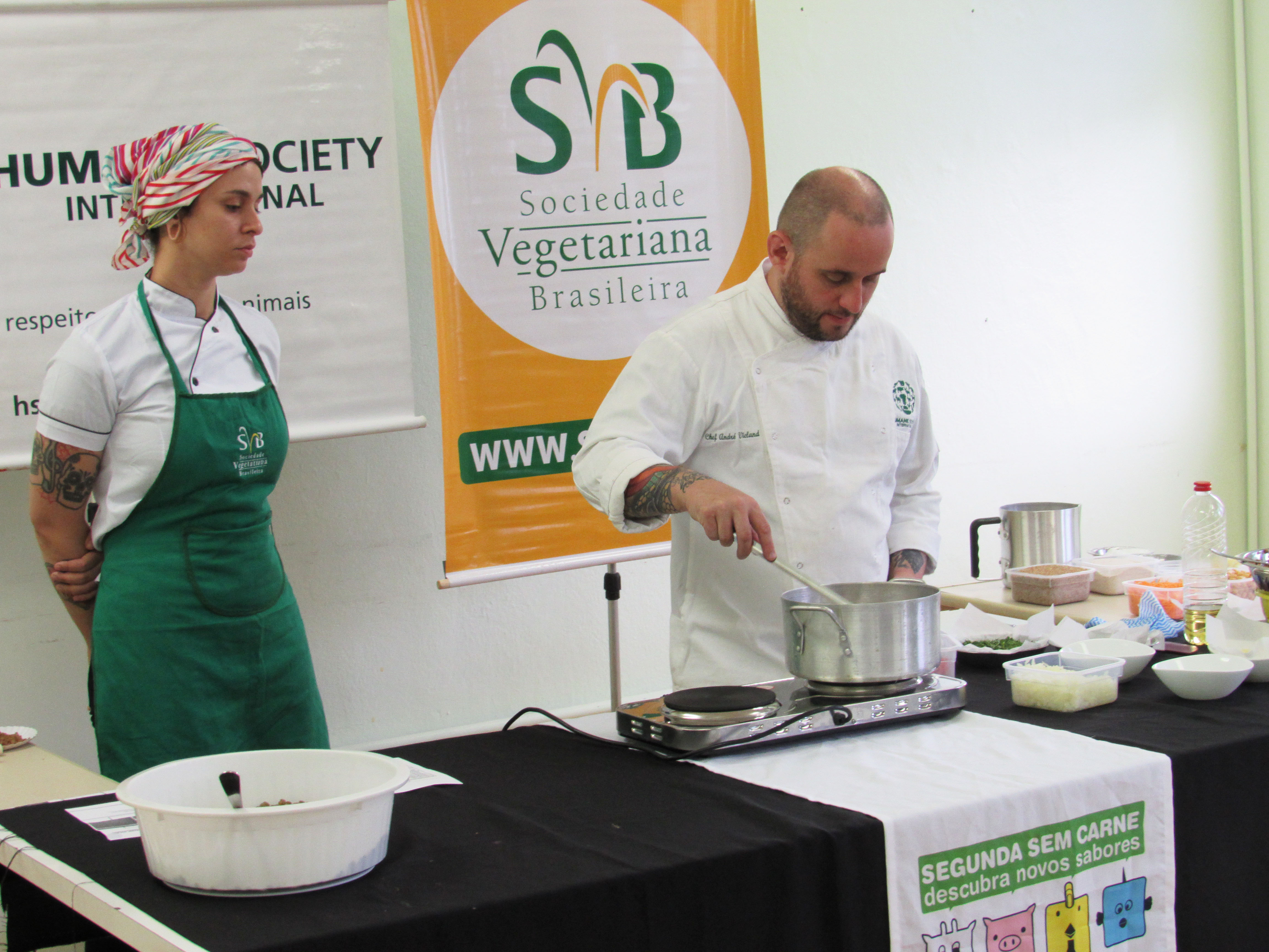 Chef de cozinha e assistente atrás de balcão com pano preto no qual estão um cooktop de duas bocas e uma penela de alumínio na qual o chef mexe a comida com uma colher metálica