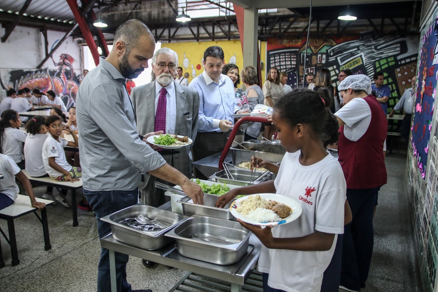Prefeito de São Paulo Bruno Covas coloca comida em prato no refeitório do serviço. Ao lado está o secretário de SMADS, Cláudio Tucci Júnior, a frente uma criança convivente e uma copeira