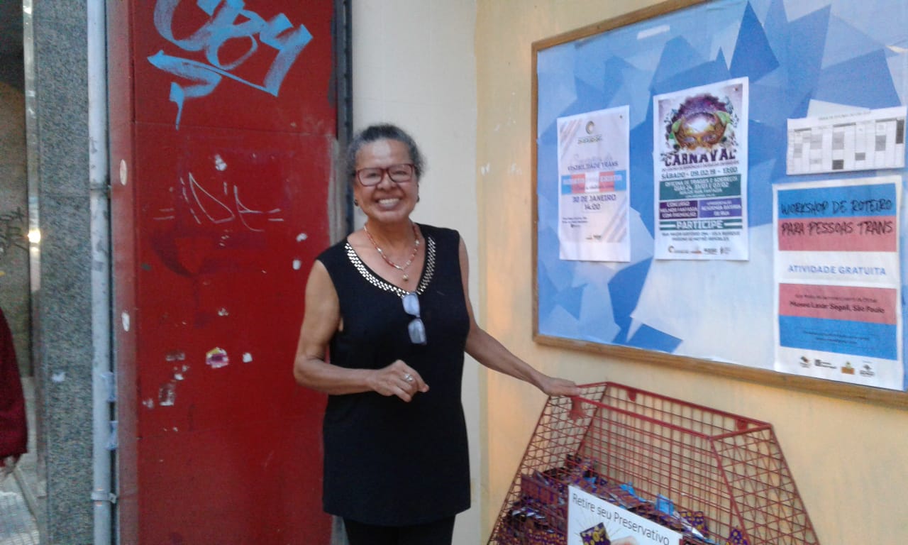 Thaís de Azevedo com vestido preto, sorrindo, em frente à porta de entrada do serviço 