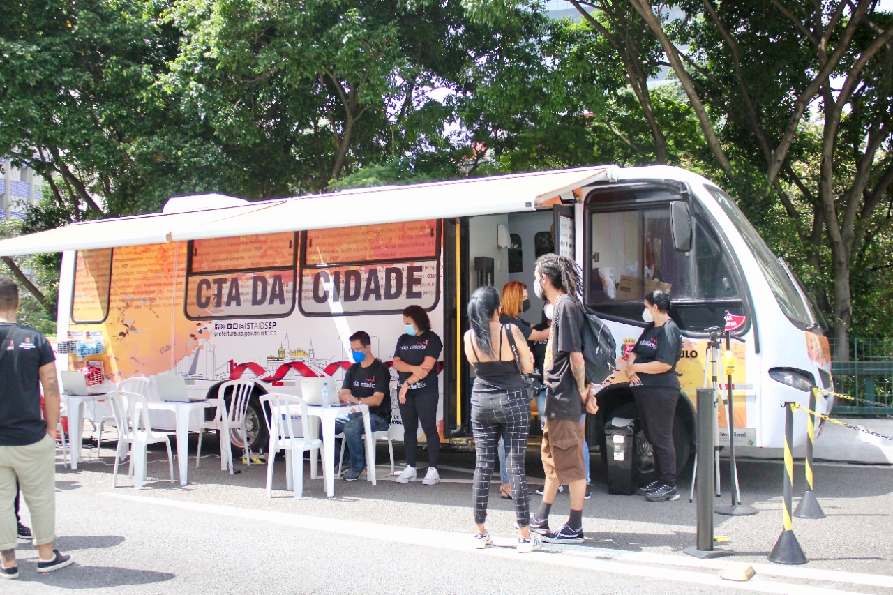 Na imagem há um ônibus laranja da CTA da cidade. Ao lado do ônibus há mesas com cadeiras e algumas pessoas em pé vestidas de preto.