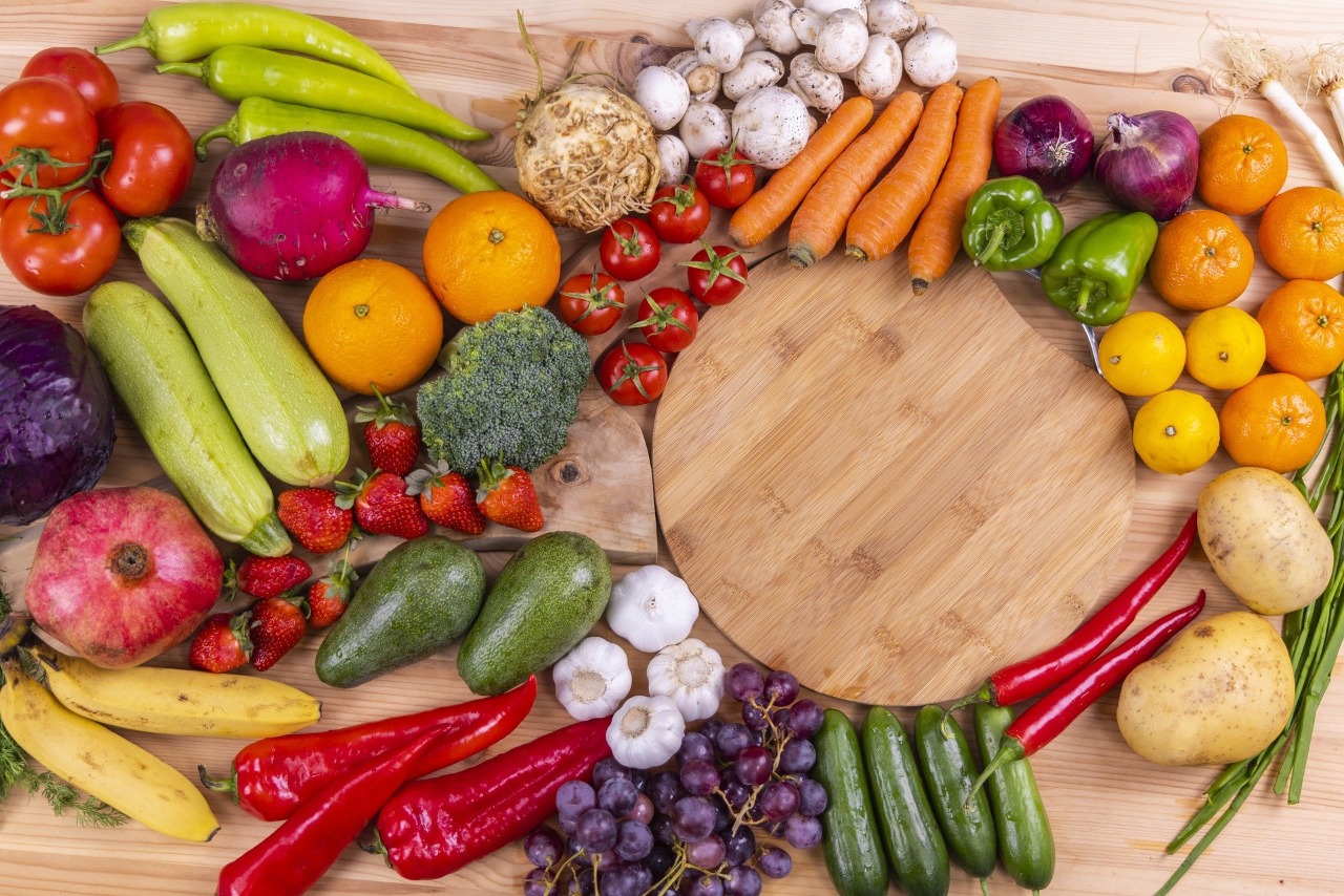 Foto de alimentos. Em cima de uma mesa de madeira há vários alimentos coloridos, dentre eles frutas, verduras e legumes. É possível ver uma tábua de madeira e ao redor dela há cenouras, tomates, pimentões, cebolas, limões, laranjas, batatas, alho, pimenta, uvas, abacates, bananas, brócolis, pitaya, morangos entre outros alimentos.