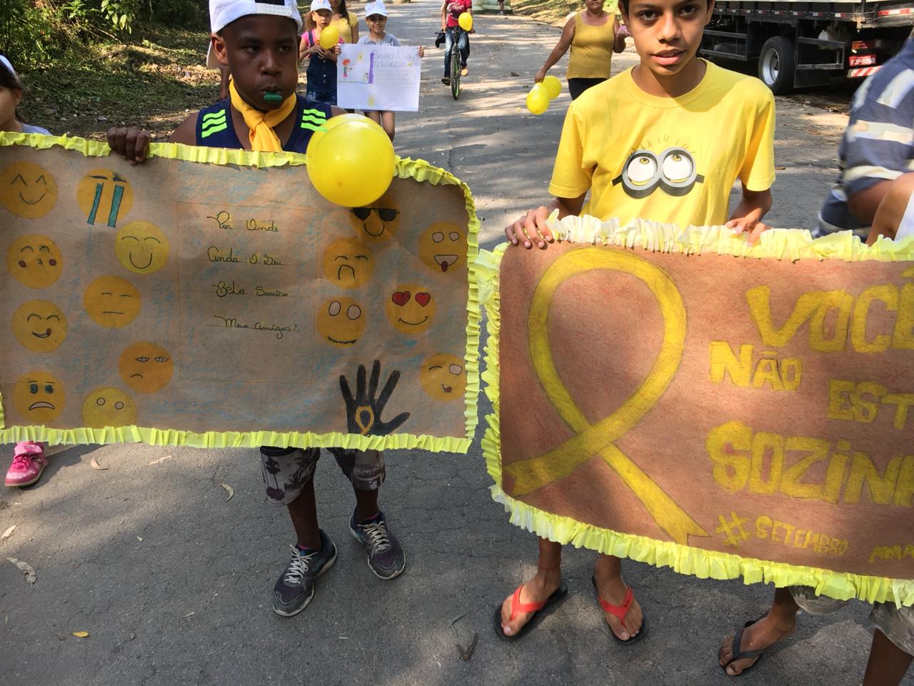 Dois meninos seguram cartazes com símbolo do setembro amarelo, desenhos de emotions e escrito “Você não está sozinho” e “Por onde anda seu belo sorriso meu amigo?” 