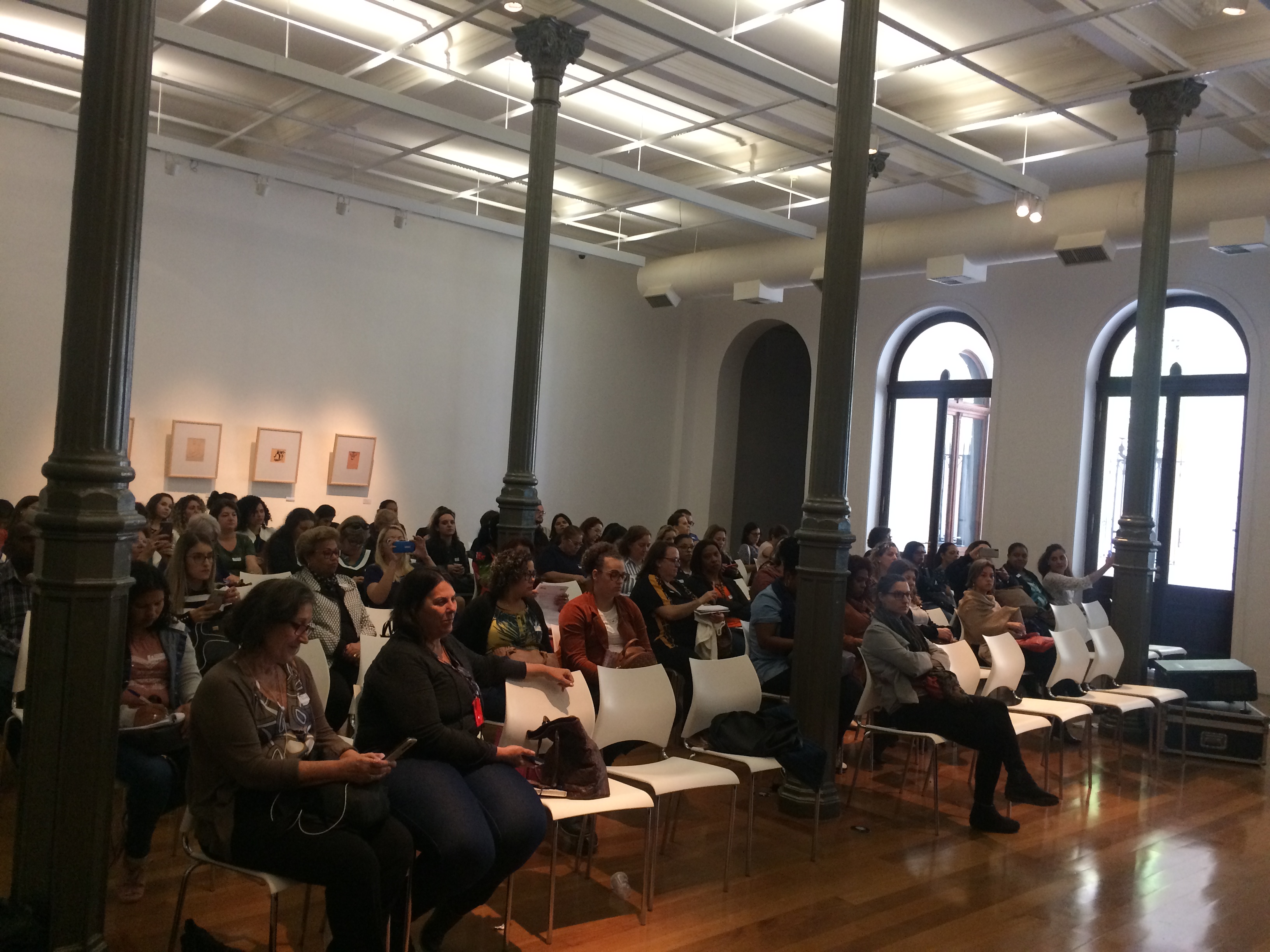 Plateia de convidados sentados em cadeiras enfileiradas em um belo auditório de fundo branco com pilastras em cinza. 