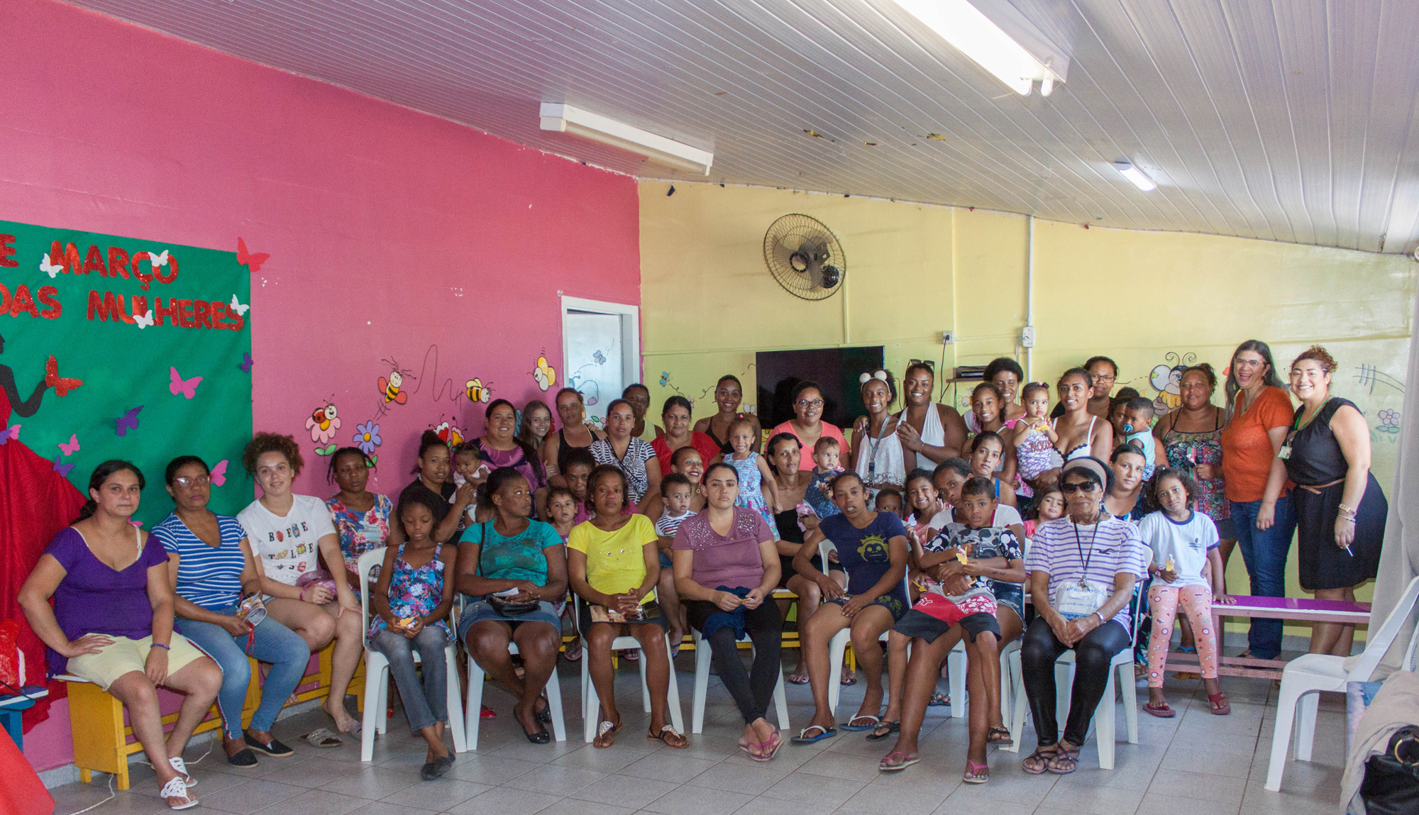 Sala onde estão reunidas, aproximadamente 40 mulheres. Algumas estão sentadas, outras em pé, olhando para a fotografia.