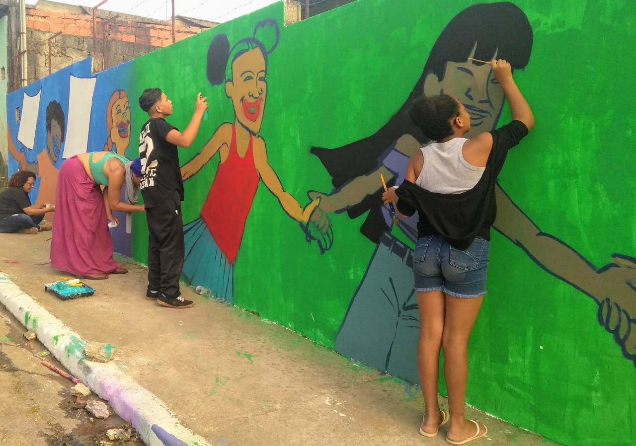 Três moças e um rapaz pintando muro do SPVV. A pintura ilustra quatro jovens de mãos dadas distribuídos pelo muro sob um fundo metade azul e metade verde com dois retângulos brancos que simbolizam o Estatuto da Criança e do Adolescente.