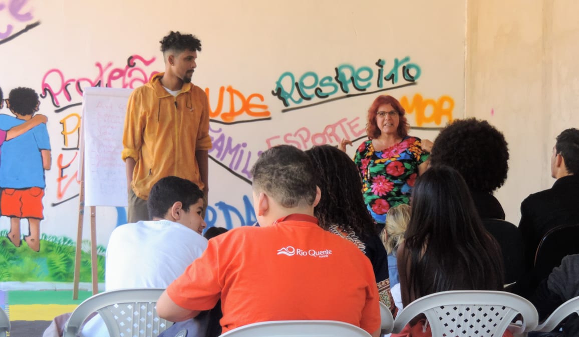 Oito jovens sentados em cadeiras de plástico assistem a palestra de uma mulher em pé com camiseta florida e cabelos curtos. Ao lado dela, um homem também em pé de moletom laranja com as mãos no bolso a ouve falar.