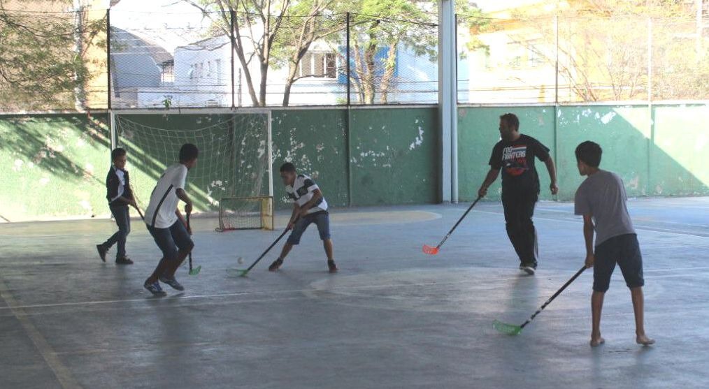 Quadra de futsal coberta com cinco pessoas ogando hóquei adaptado. Em suas mãos estão bastões de metal. 