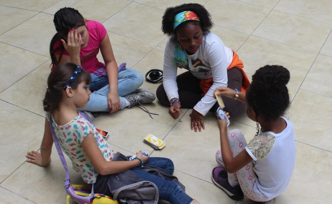 Quatro crianças sentadas no chão em roda jogando uno, um jogo de cartas. 