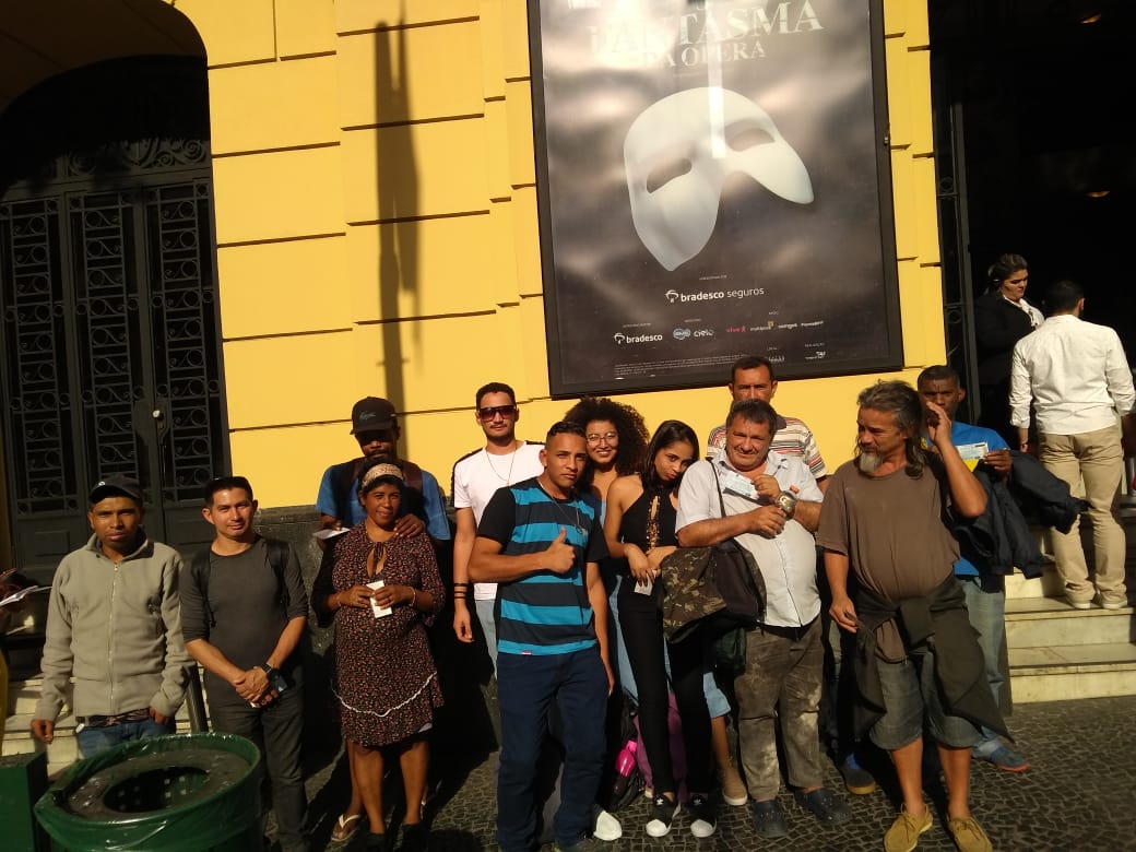 Doze pessoas em pé posando para a fotografia. Elas estão em frente à parede amarela do Teatro Renault, que exibe um grande cartaz em preto com a figura de uma máscara branca, e escrito ‘O Fantasma da Ópera’. 