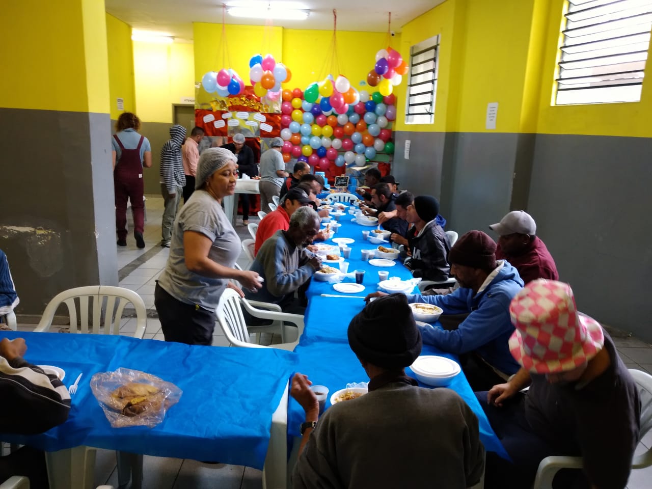 Aproximadamente quinze pessoas sentadas em mesas se alimentando. De fundo, painel de bexigas coloridas e mesa de aniversário enfeitada com bolo decorado, toalhas azuis, e pequena lousa quadrada onde está escrito “Feliz Aniversário”. 