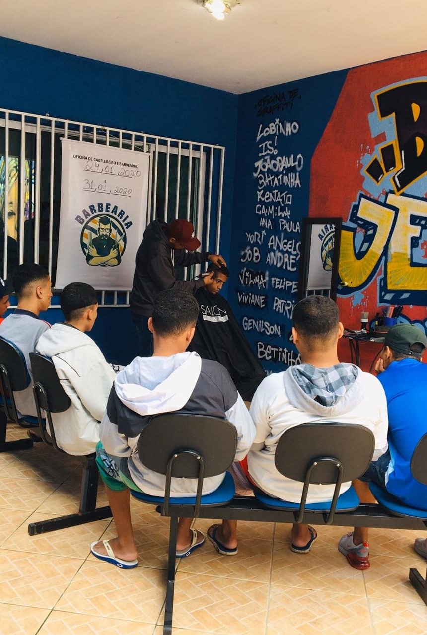 Seis garotos aparecem sentados de costas. Eles observam ao professor que aparece cortando o cabelo de garoto sentado. 