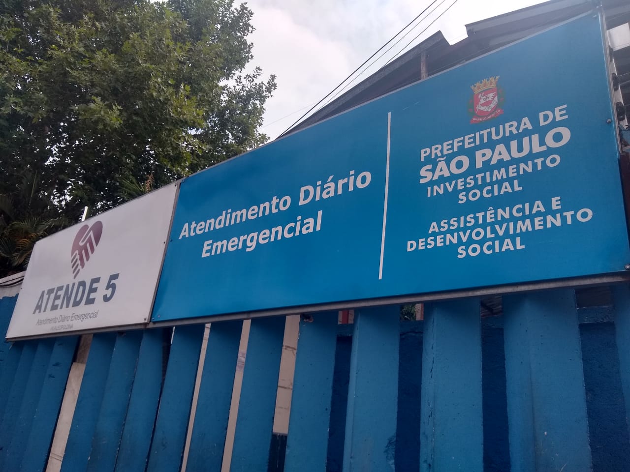 Imagem da placa do serviço na parte externa do local. A placa azul contem as seguintes informações: Atendimento Diário Emergencial – Vila Leopoldina e o logo da Prefeitura da cidade de São Paulo, logo embaixo, Assistência e Desenvolvimento Social. 