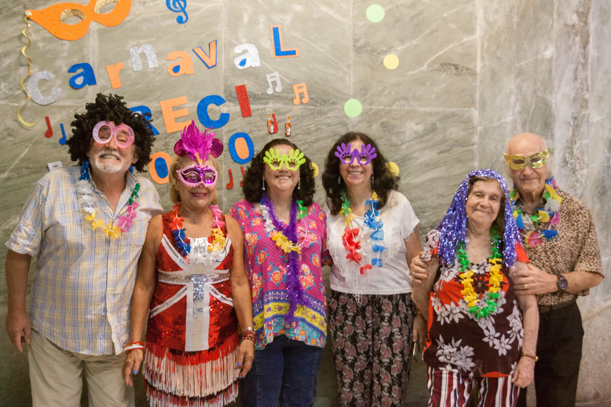 Seis idosos posam sorrindo para a foto. Eles usam apetrechos coloridos de carnaval. No fundo, uma parede onde se lê “Carnaval CRECI 2020” com letras e símbolos coloridos em volta.