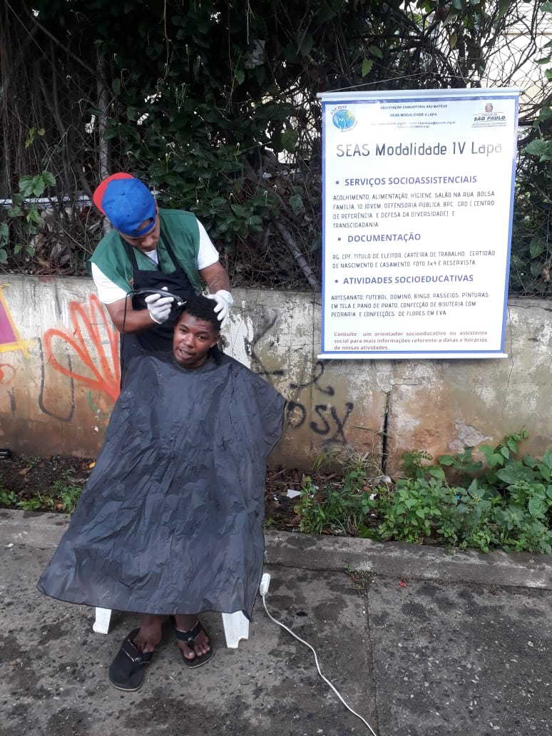 Homem sentado em cadeira enquanto assistente social corta seu cabelo. De fundo, um banner que leva o nome do serviço e suas ações. 