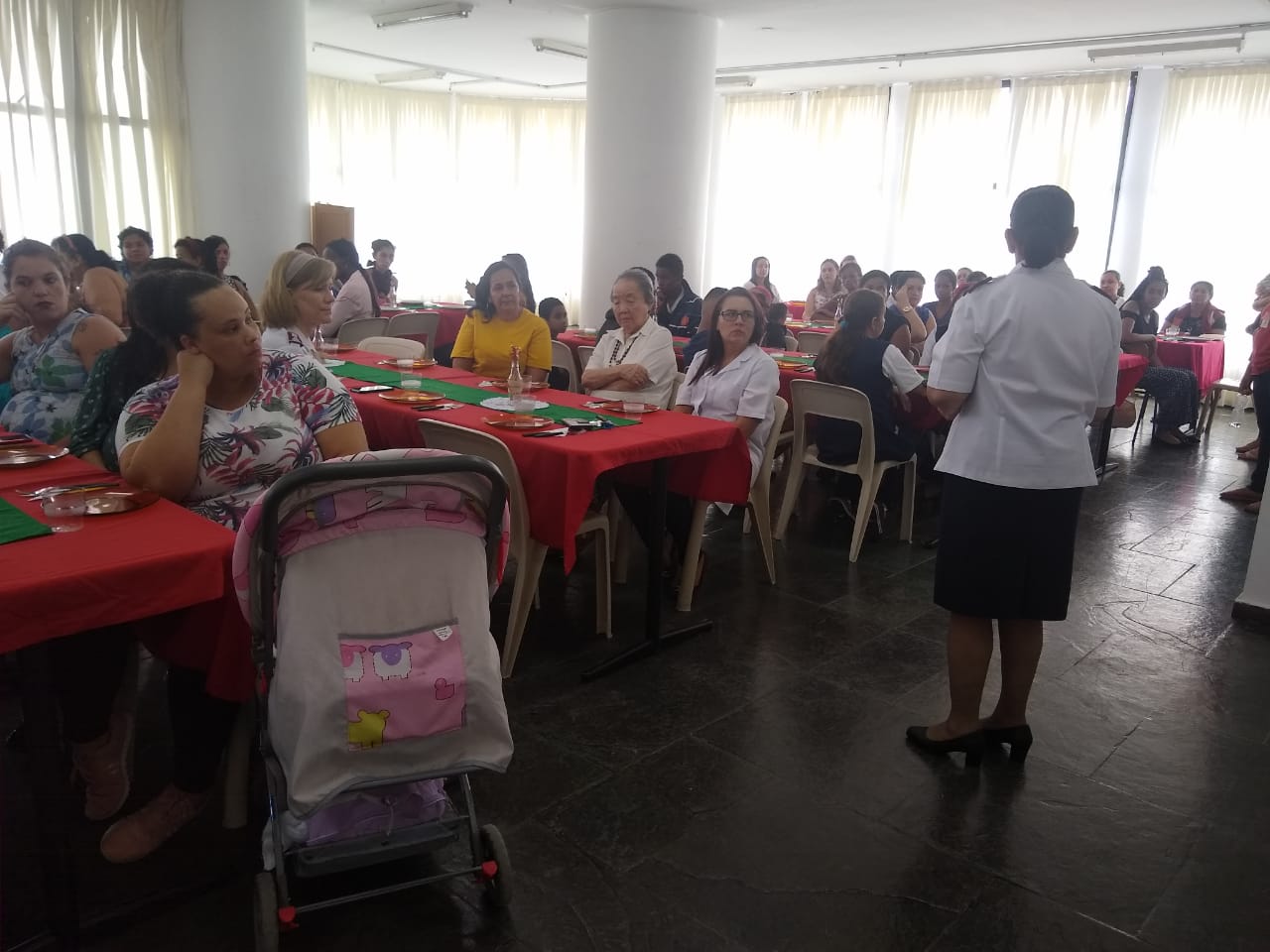 Mulheres reunidas em salão para almoço de confraternização. Elas observam, sentadas, Major Jurema se apresentando a frente do salão.