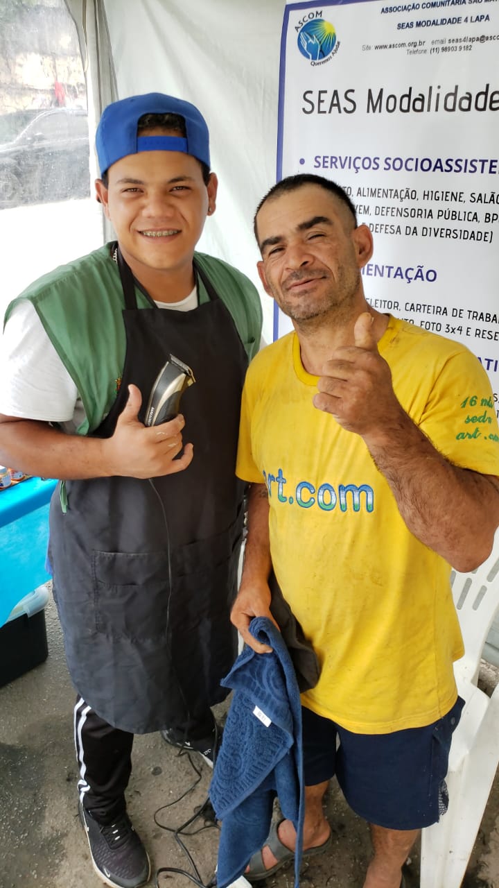 Homem atendido aparece de cabelo e barba cortada ao lado do assistente que o atendeu.