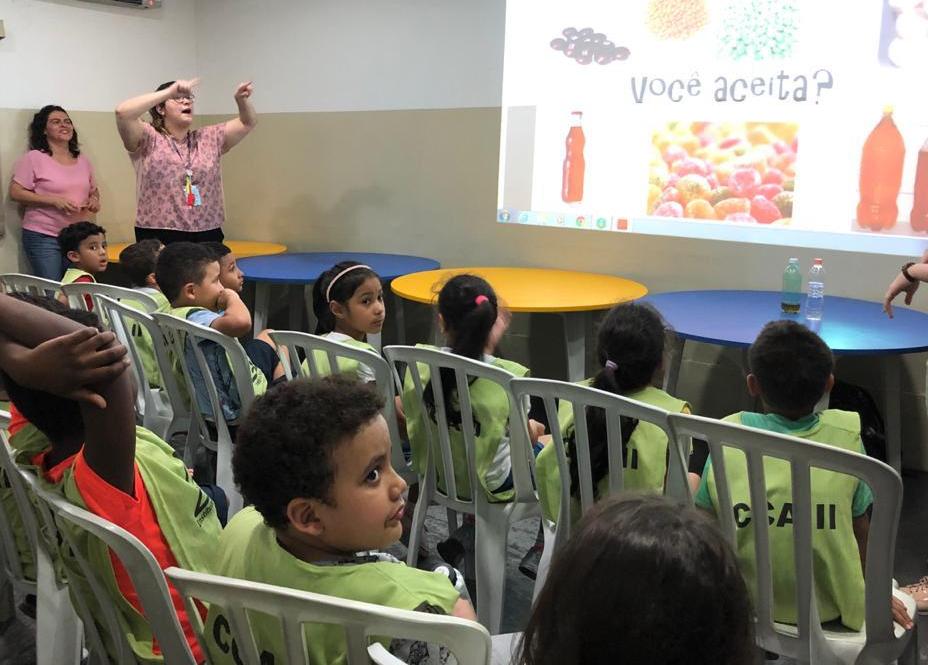 Crianças reunidas em sala aparecem assistindo palestra. À frente da sala, telão que exibe slides educativos sobre o tema. 