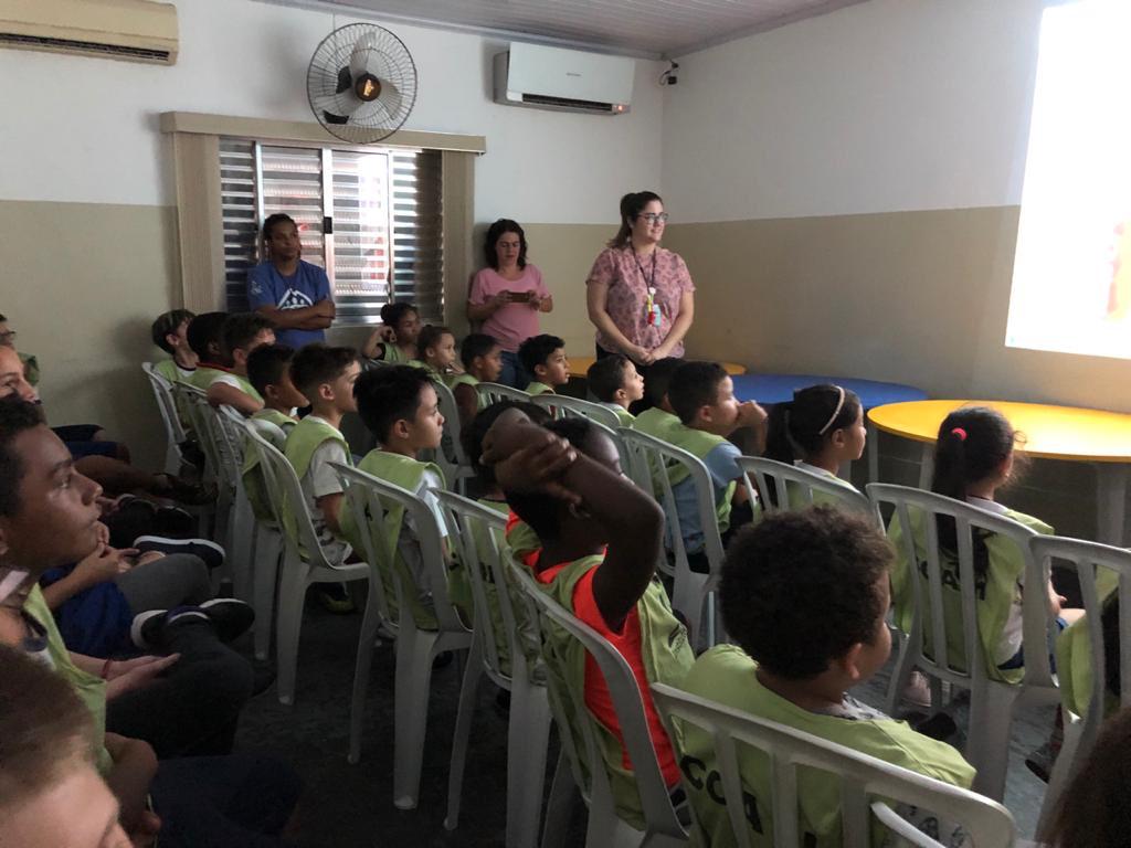 36 crianças aparecem em uma sala, sentadas em cadeiras plásticas enfileiradas, assistindo a vídeo educativo de orientação exibido em telão. 