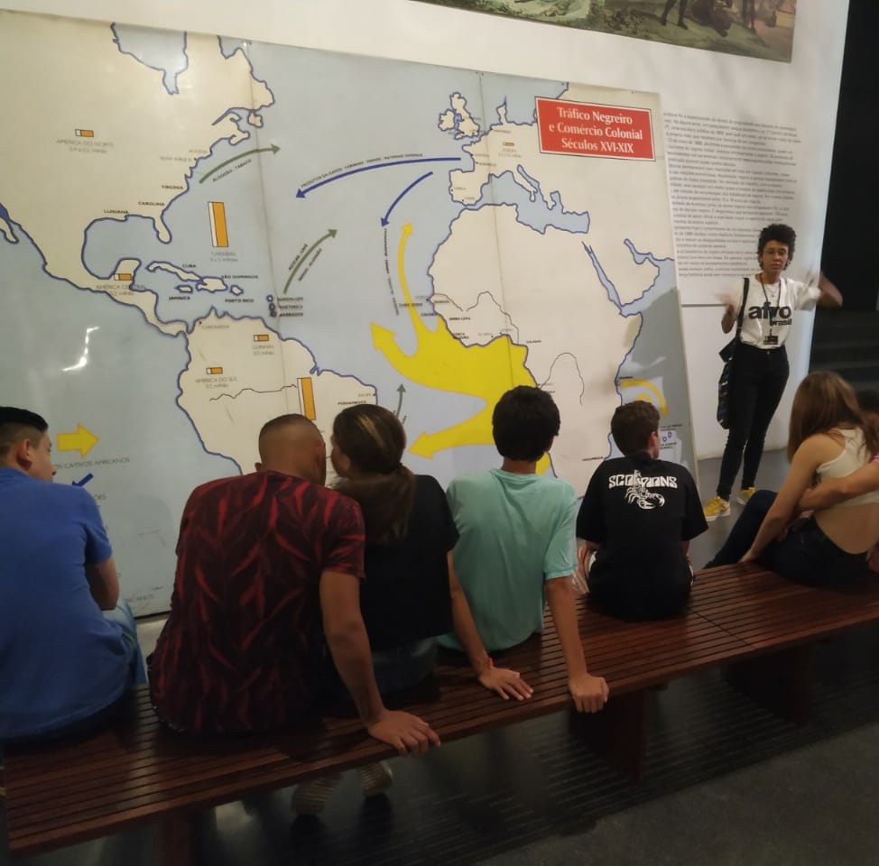 Seis visitantes do Museu aparecem de costas, sentados em banco de madeira ouvindo a explicação de monitora do Museu que está de pé e gesticula ao lado de grande mapa com o seguinte título “Tráfico Negreiro e Comércio Colonial – Séculos XVI – XIX”. 