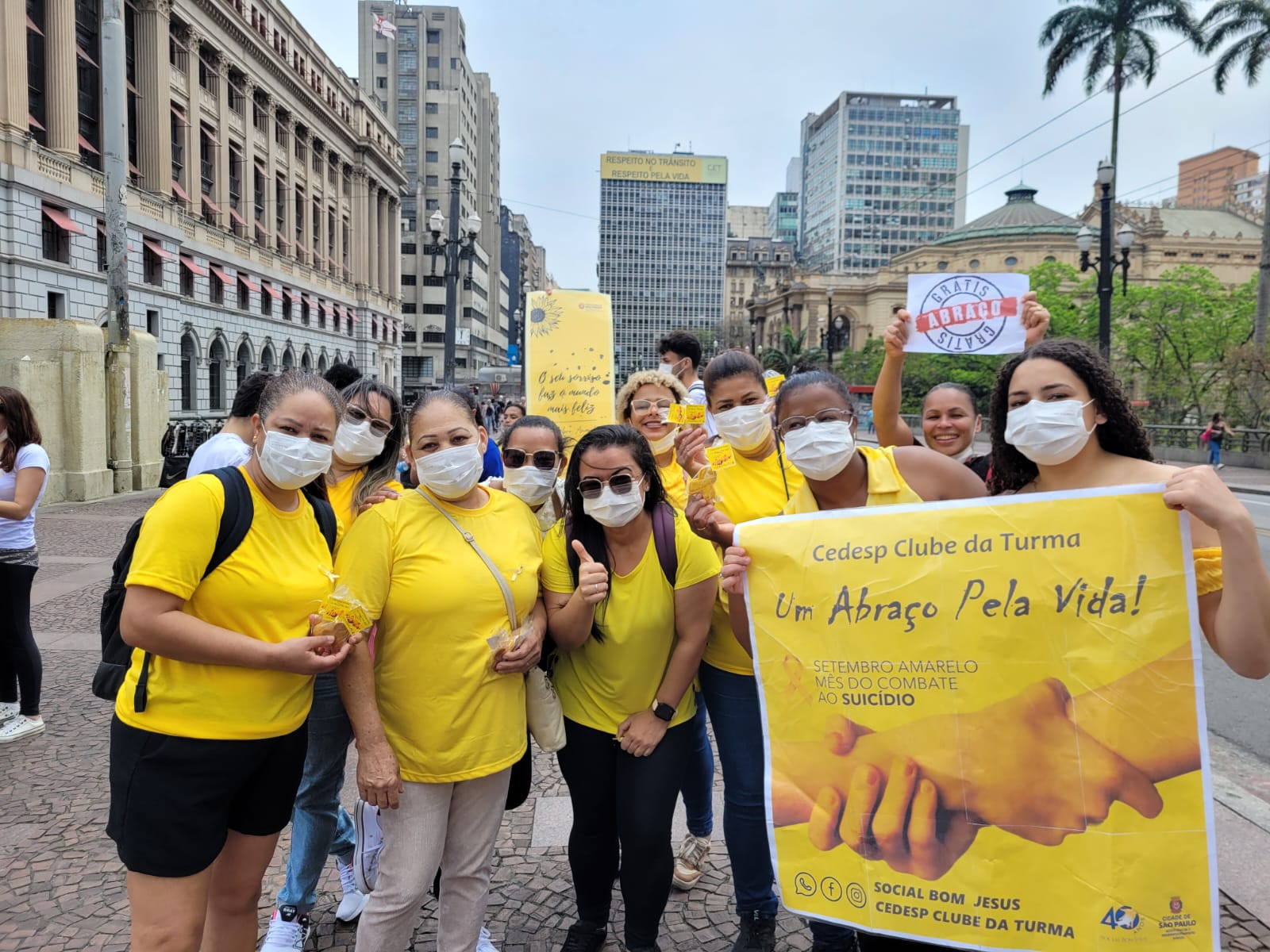Na imagem mostram um grupo de pessoas vestidas de camisetas amarelas, sorrindo para a câmera, em meio aos prédios da cidade de São Paulo. Algumas pessoas seguram cartazes escritos "CEDESP Clube da Turma" e " Um abraço pela vida".