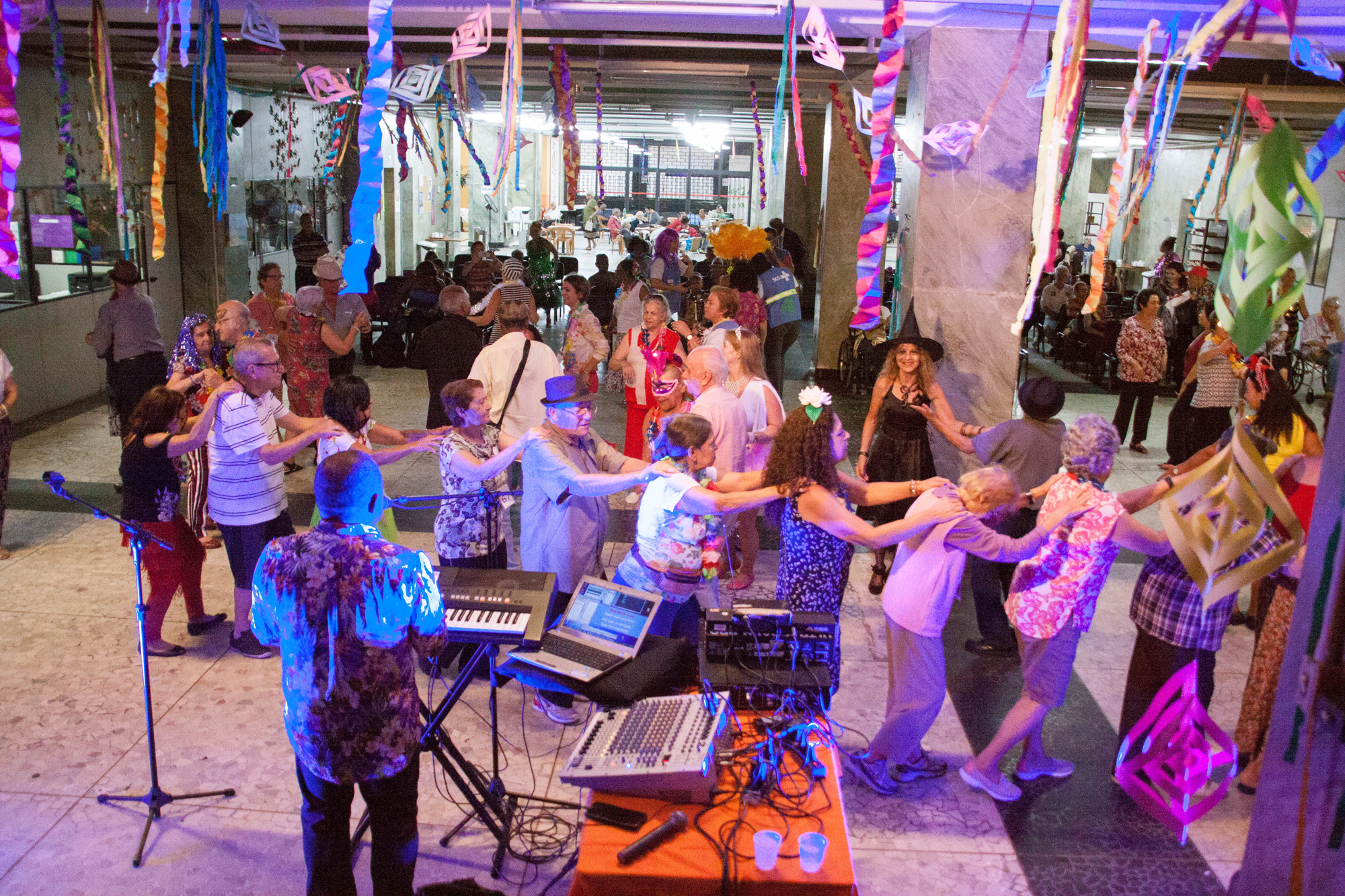 Grande salão decorado com enfeites e luzes coloridas. Nele, aparecem dezenas de idosos dançando, músico tocando. Cadeiras no fundo do salão, onde alguns idosos se encontram sentados repousando e observando a festa.