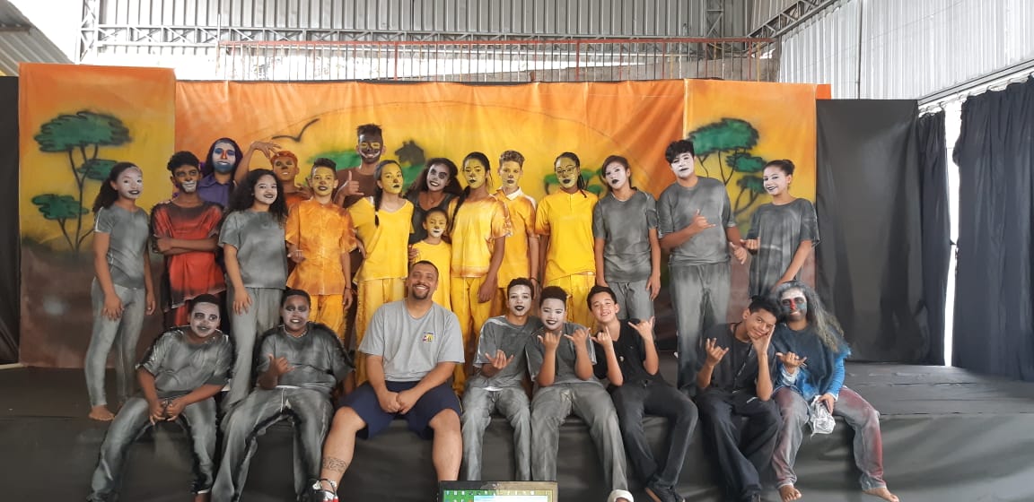 Aproximadamente trinta pessoas, sendo crianças e adolescentes e professor no palco, posando para fotografia. A maioria está caracterizada como personagens da peça Rei Leão, com seus rostos pintados, adereços na cabeça, e trajes coloridos.