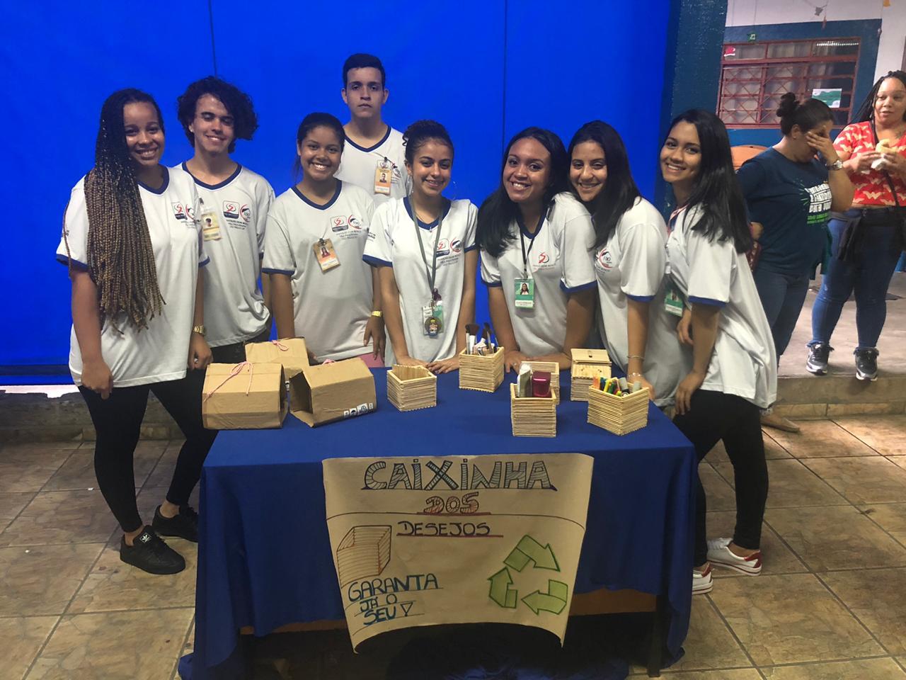 Oito alunos sorrindo para a foto atrás de mesa que expõe projeto educacional desenvolvido pelos mesmos. 