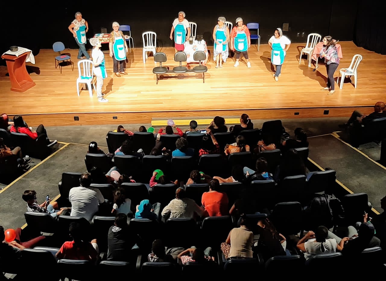 Auditório onde estão espectadores em poltronas assistindo a peça encenada pelos conviventes no palco. 