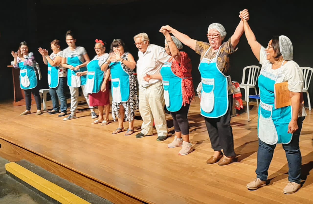 Atores no palco de mãos dadas agradecendo aos aplausos do público no final da peça e se despedindo.