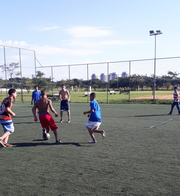 Seis jovens estão jogando futebol sob um gramado sintético. No centro da imagem há um garoto com a bola nos pés rodeado de outros jogadores. Ao fundo, o restante do Parque Villa-Lobos e prédios atrás.
